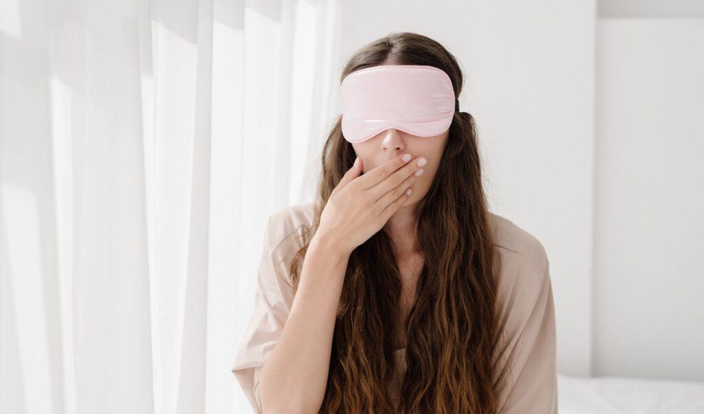 Woman with eye mask, yawning, sitting on bed