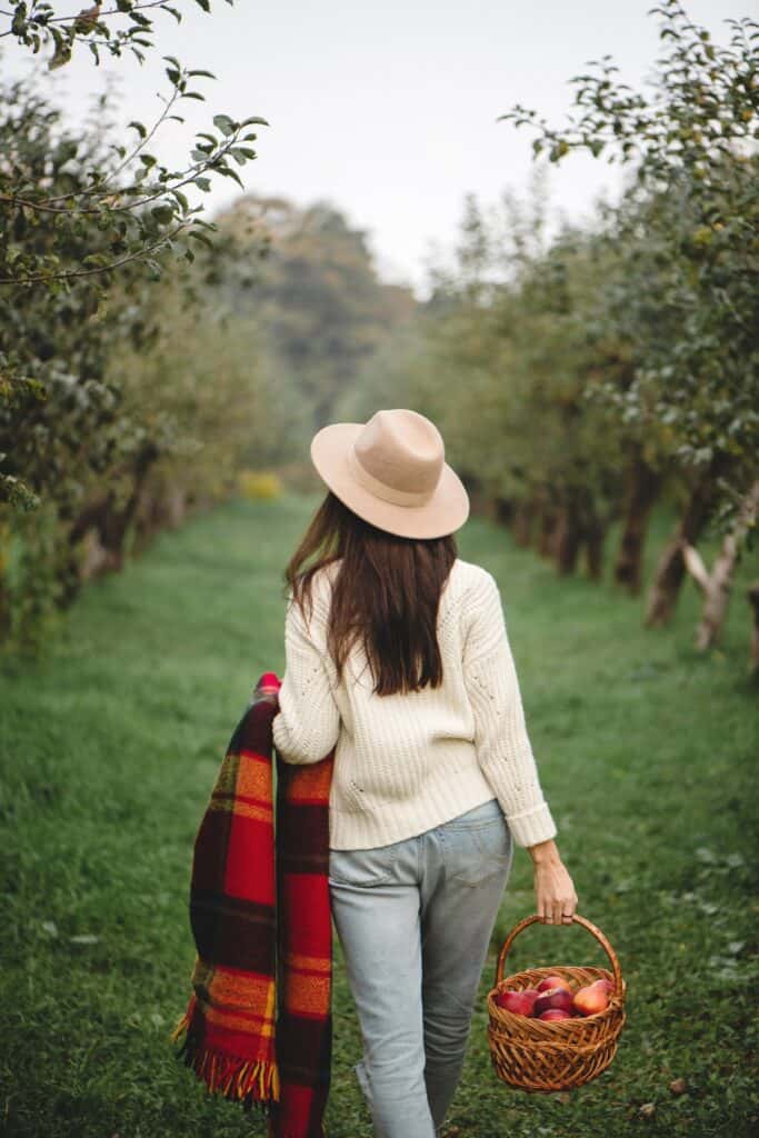 Woman wearing a hat walking in an orchard holding a checked blanket and basket of food